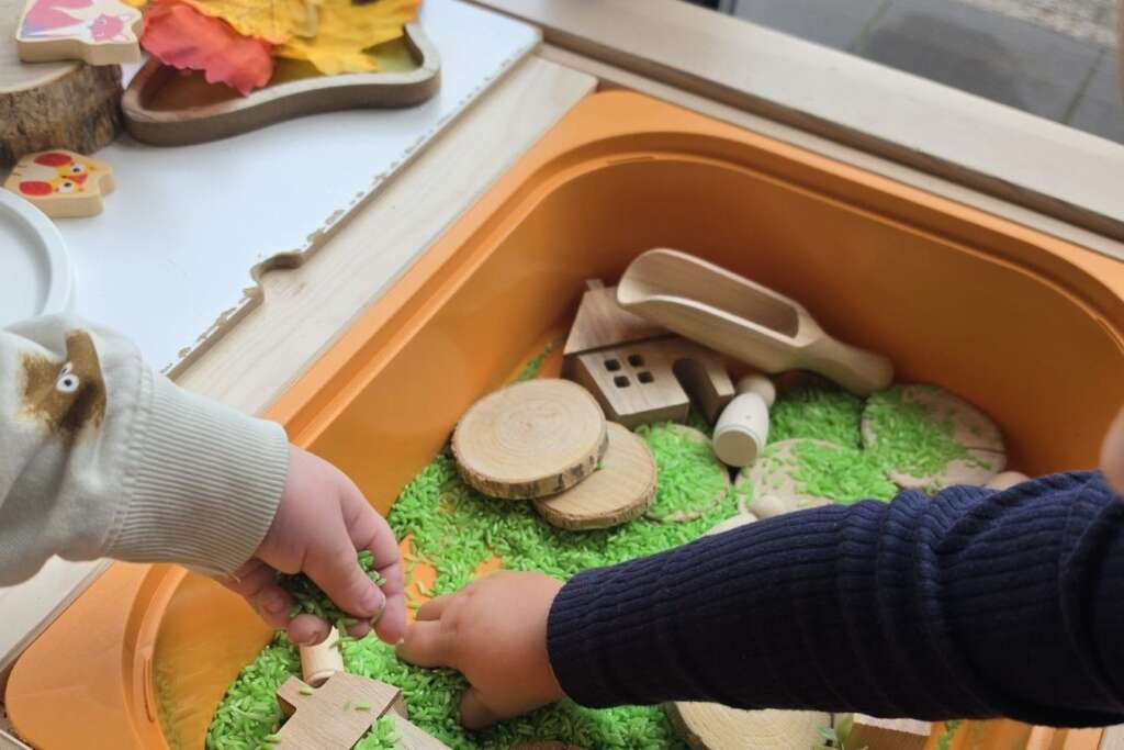 Twee peuters spelen met groene rijst en houten huisjes in een speelbak